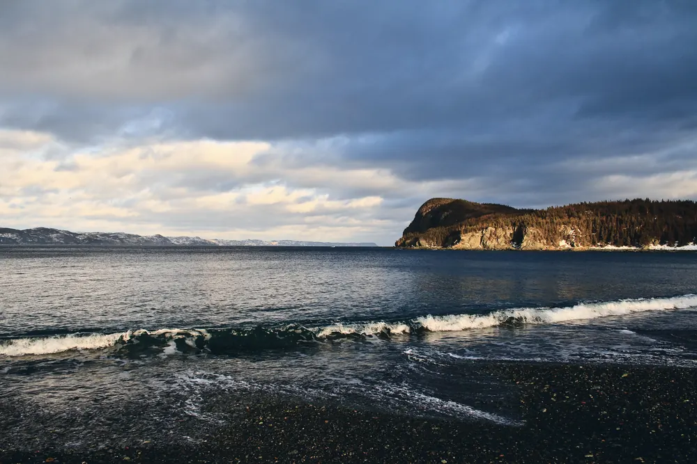 A wave breaking along a stony beach at Harry's Harbour, low winter sun lighting the headland across the bay.