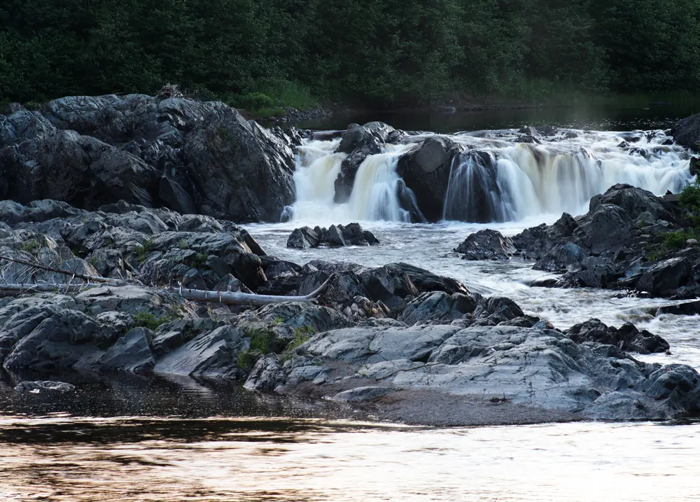 Indian River pouring white over a ledge of dark rock and running on between the stones, forest behind.
